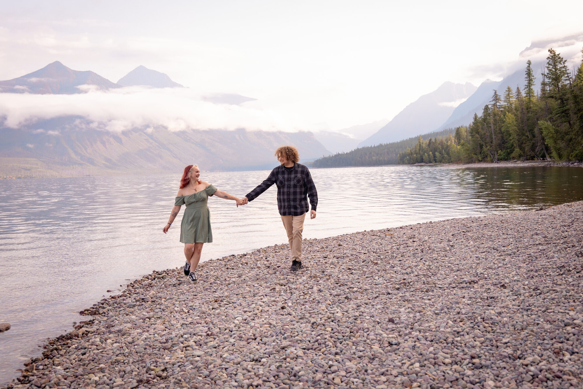 Couple walking hand in hand along a rocky lakeshore beach with mountains in the mist
