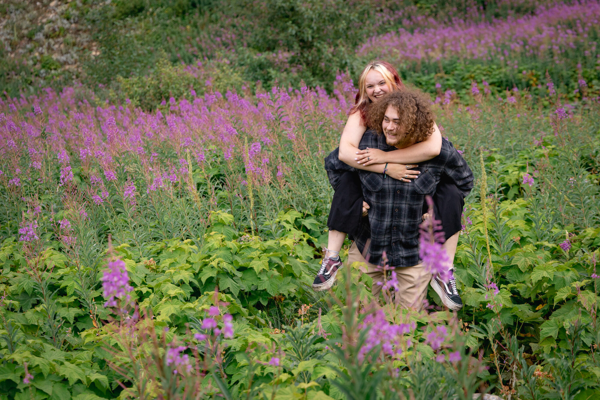 Couple laughing during a piggyback ride through a field of fireweed wildflowers